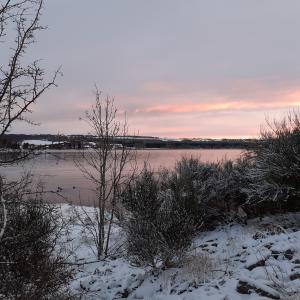 snowy scene of water with hills in distance, pink setting sun reflected on water
